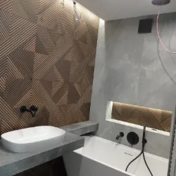 Nowoczesna łazienka z drewnianą ścianą o geometrycznym wzorze, białą umywalką nablatową na szarym blacie, białą wanną i szarą ścianą z wnęką podświetloną drewnem.