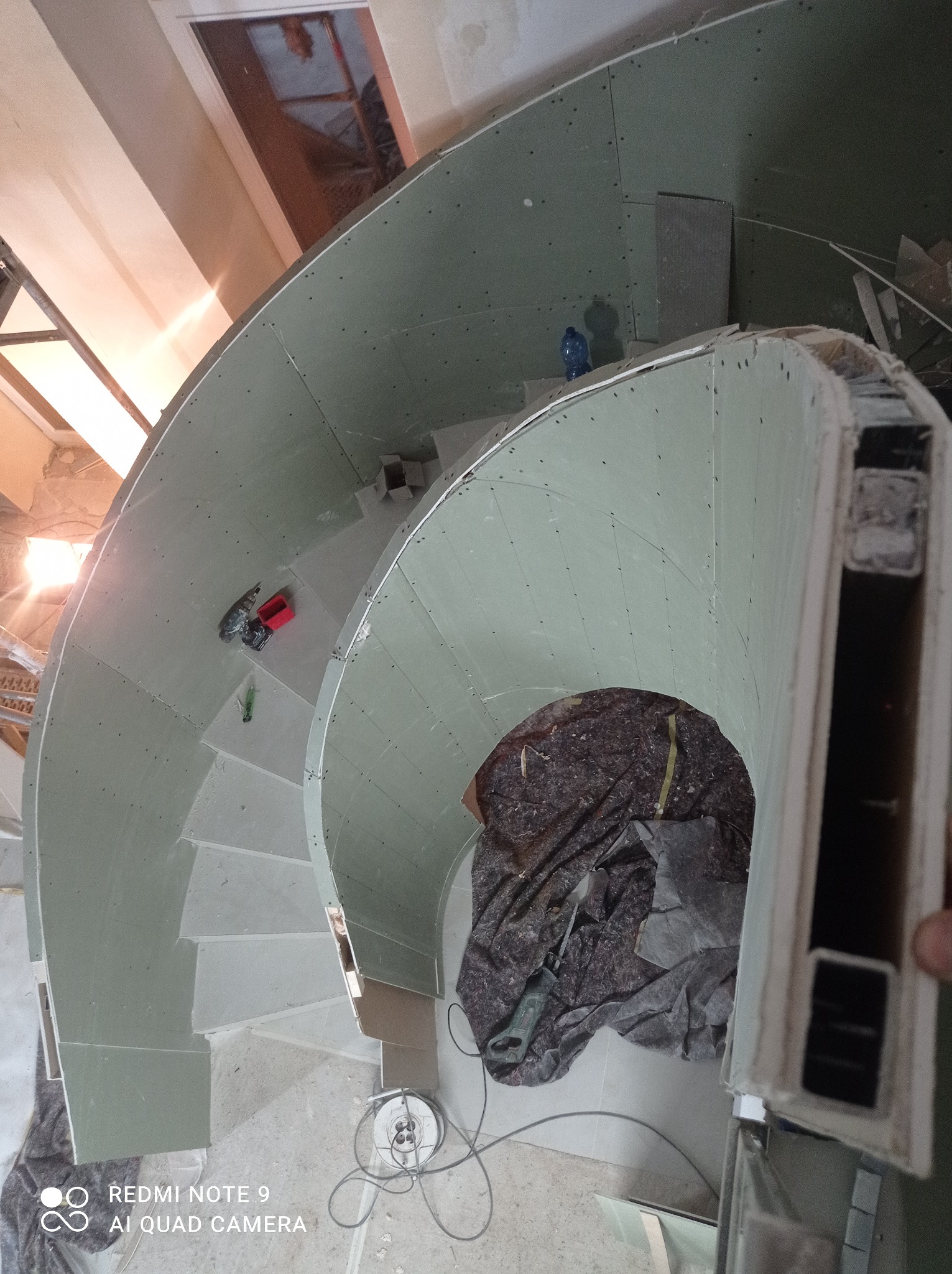 Spiralne schody w trakcie obudowy płytami kartonowo-gipsowymi, widok z góry, z widocznymi narzędziami i fragmentami konstrukcji.