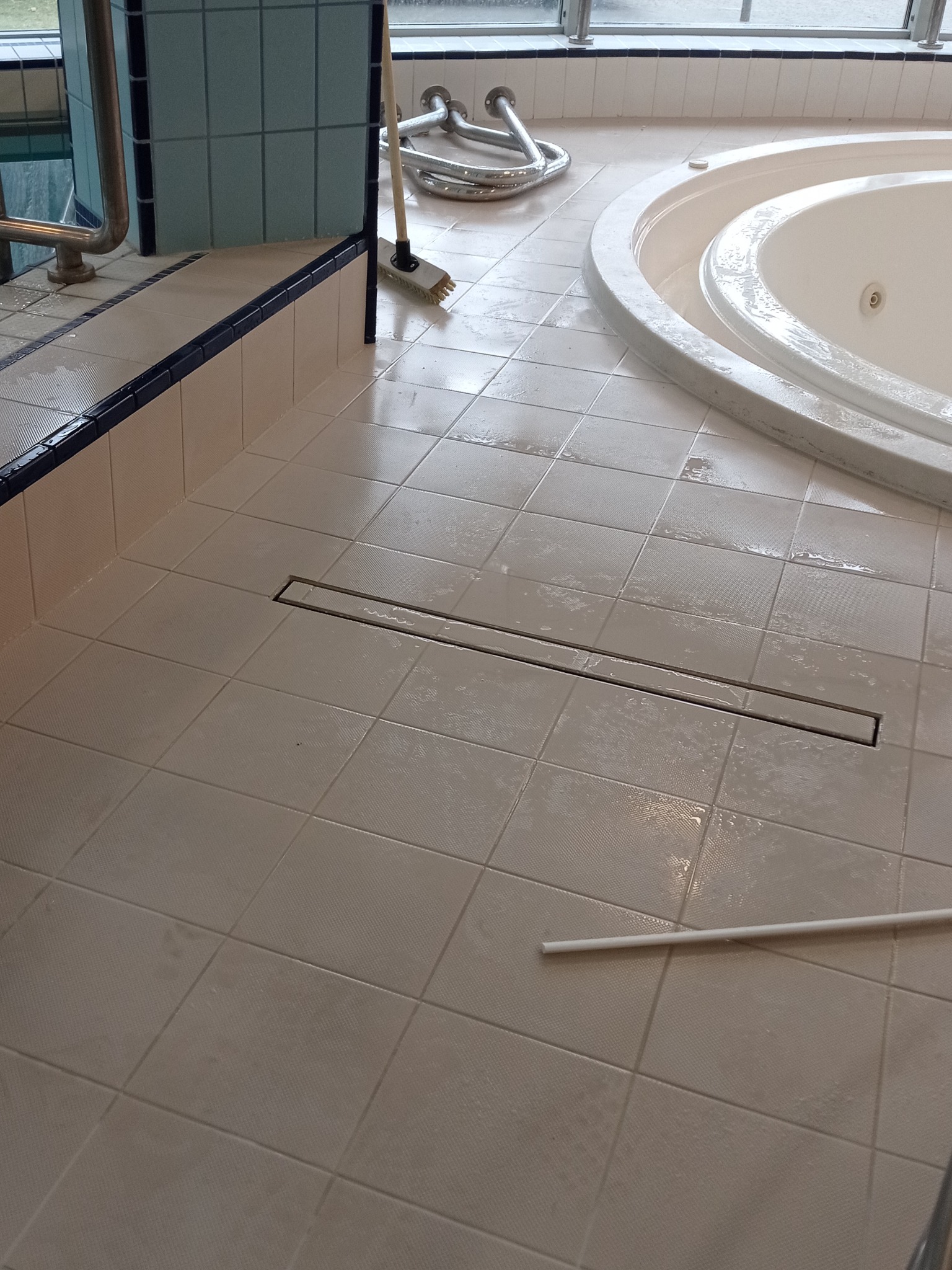 Jasne płytki podłogowe po ułożeniu wokół wanny z odpływem liniowym. Widoczne akcesoria montażowe i szczotka. Minimalistyczny design.