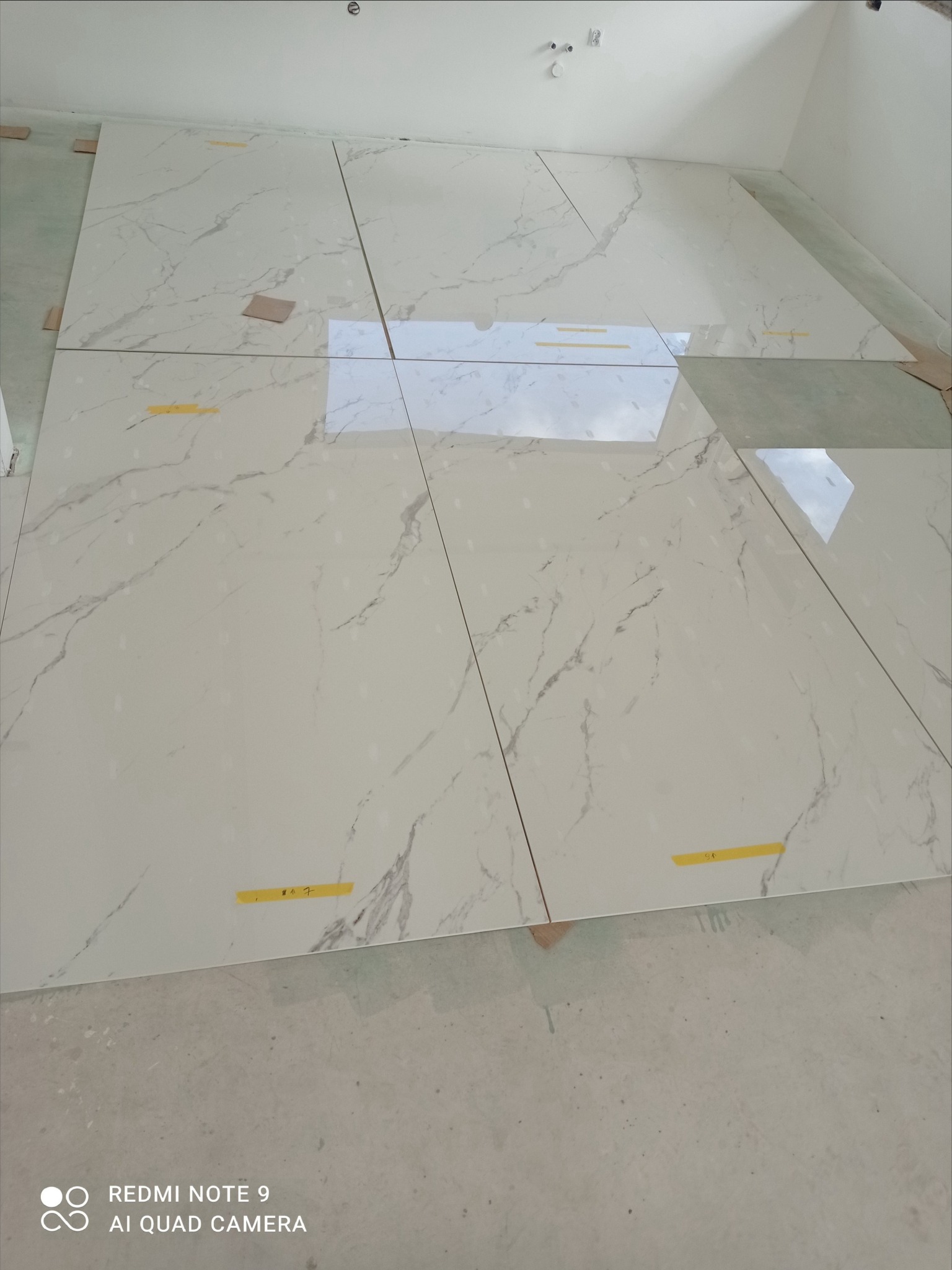 Układanie jasnych, marmurkowych płytek podłogowych w nowym wnętrzu. Widoczne żółte taśmy i drewniane kliny dystansowe. Odbicie światła w polerowanej powierzchni.