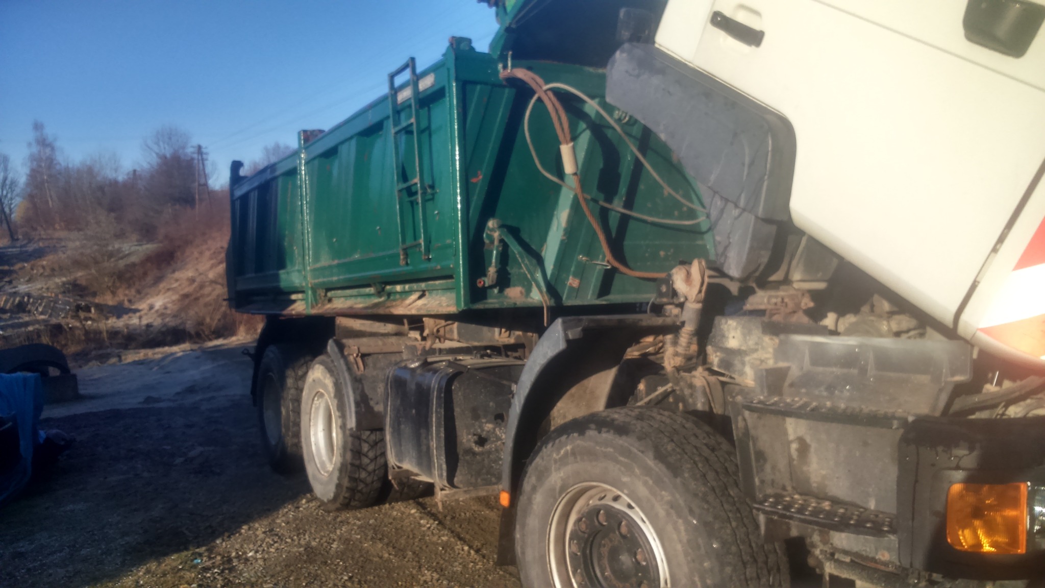 Zielona ciężarówka wywrotka z podniesioną kabiną na żwirowym podłożu, widoczne drzewa w tle.