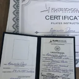 Certyfikat instruktora Pilates, otwarty dokument legitymacji z danymi osobowymi i pieczątkami, leżący na drewnianym podłożu, z kilkoma dodatkowymi certyfikatami w tle.