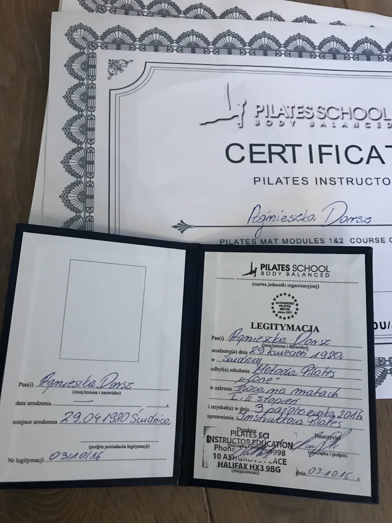 Certyfikat instruktora Pilates, otwarty dokument legitymacji z danymi osobowymi i pieczątkami, leżący na drewnianym podłożu, z kilkoma dodatkowymi certyfikatami w tle.