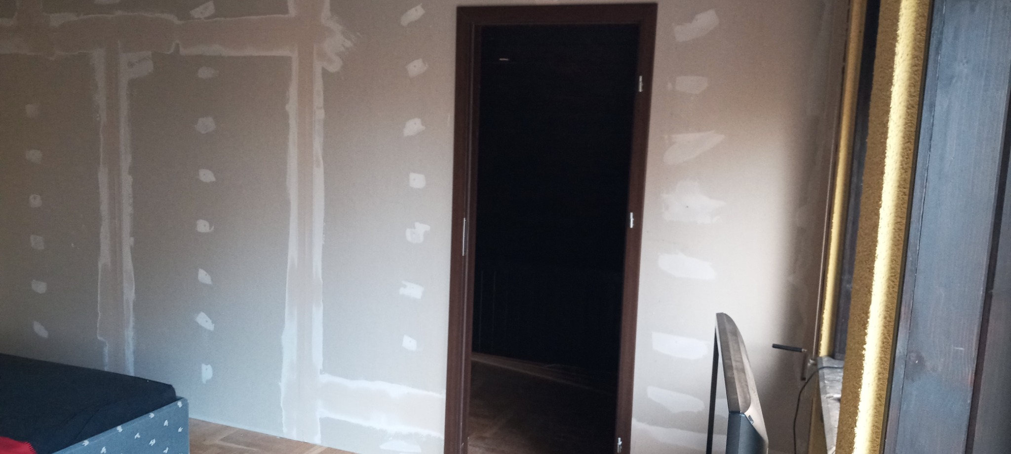 Ściana z płyt gipsowo-kartonowych przed malowaniem, widoczne łączenia wypełnione szpachlą, w tle zamontowane ciemne drzwi z brązową ościeżnicą.