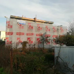 Budynek mieszkalny w trakcie prac elewacyjnych, osłonięty siatką z nadrukowanym logo firmy FAST i DAN-MET, widok zza ogrodzenia i zieleni.