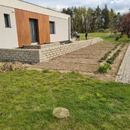 Wznoszenie muru oporowego z jasnych bloczków przy nowoczesnym domu z drewnianą elewacją; w tle widoczni dwaj pracownicy i betoniarka.