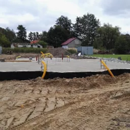 Wylana płyta fundamentowa z izolacją pionową, wystającymi prętami zbrojeniowymi i żółtymi rurami instalacyjnymi, otoczona piaskiem z widocznymi śladami opon, w tle zabudowa jednorodzinna.