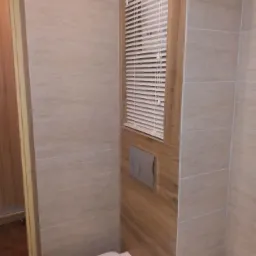 Wykończona łazienka z beżowymi płytkami imitującymi kamień, zabudowaną spłuczką WC i okienkiem z żaluzjami.