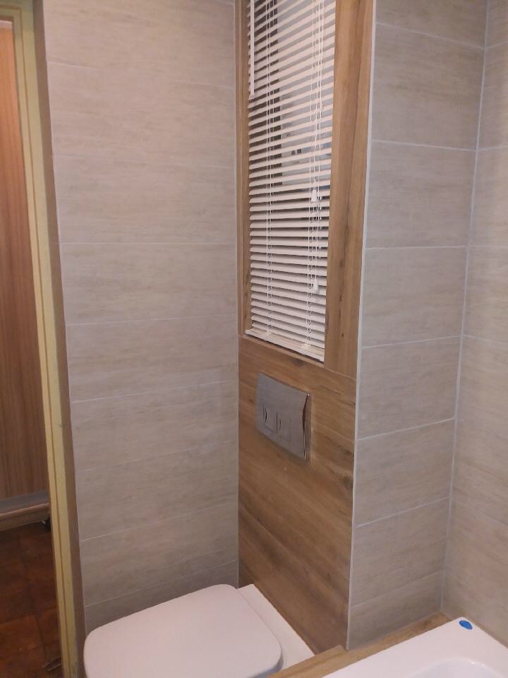 Wykończona łazienka z beżowymi płytkami imitującymi kamień, zabudowaną spłuczką WC i okienkiem z żaluzjami.