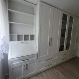 Biała zabudowa meblowa z biurkiem sekretarzykiem, p&oacute;łkami i szafami z przeszklonymi drzwiami, czarne uchwyty, panele podłogowe.