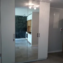 Biała szafa z lustrzanymi drzwiami w przedpokoju, widok na kuchnię z szarym tłem i drewnianymi szafkami, nowoczesne oświetlenie sufitowe.