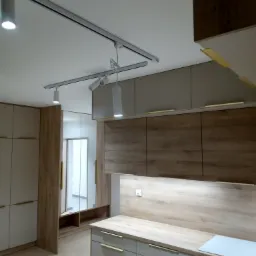 Nowoczesna kuchnia z drewnianymi akcentami, szare szafki z poziomymi, złotymi uchwytami, oświetlenie szynowe i blat w odcieniach drewna.