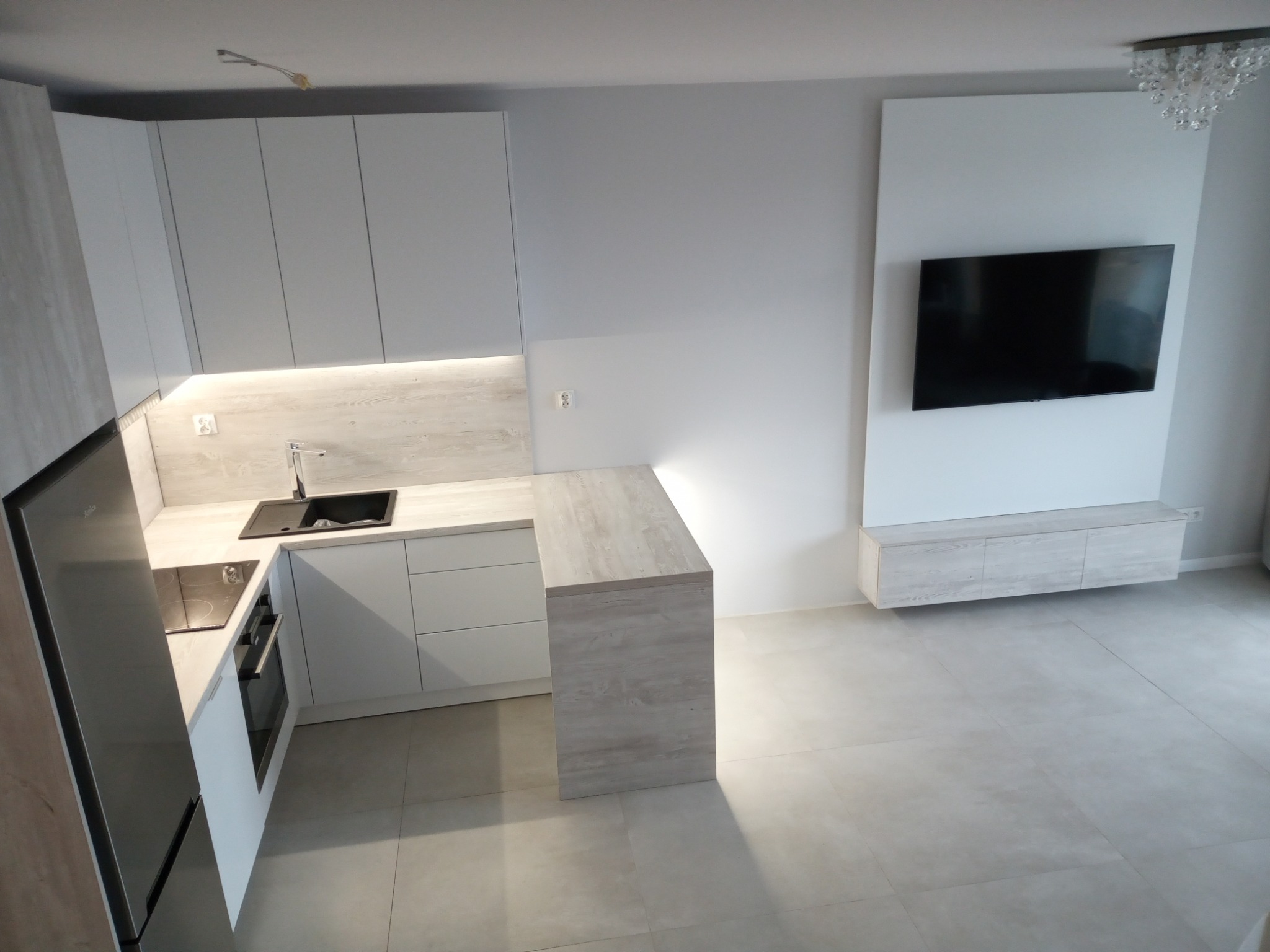 Nowoczesna, minimalistyczna kuchnia z białymi frontami szafek i blatem w odcieniach jasnego drewna, czarnym zlewem i płytą indukcyjną, z wyspą kuchenną i telewizorem na ścianie w tle.