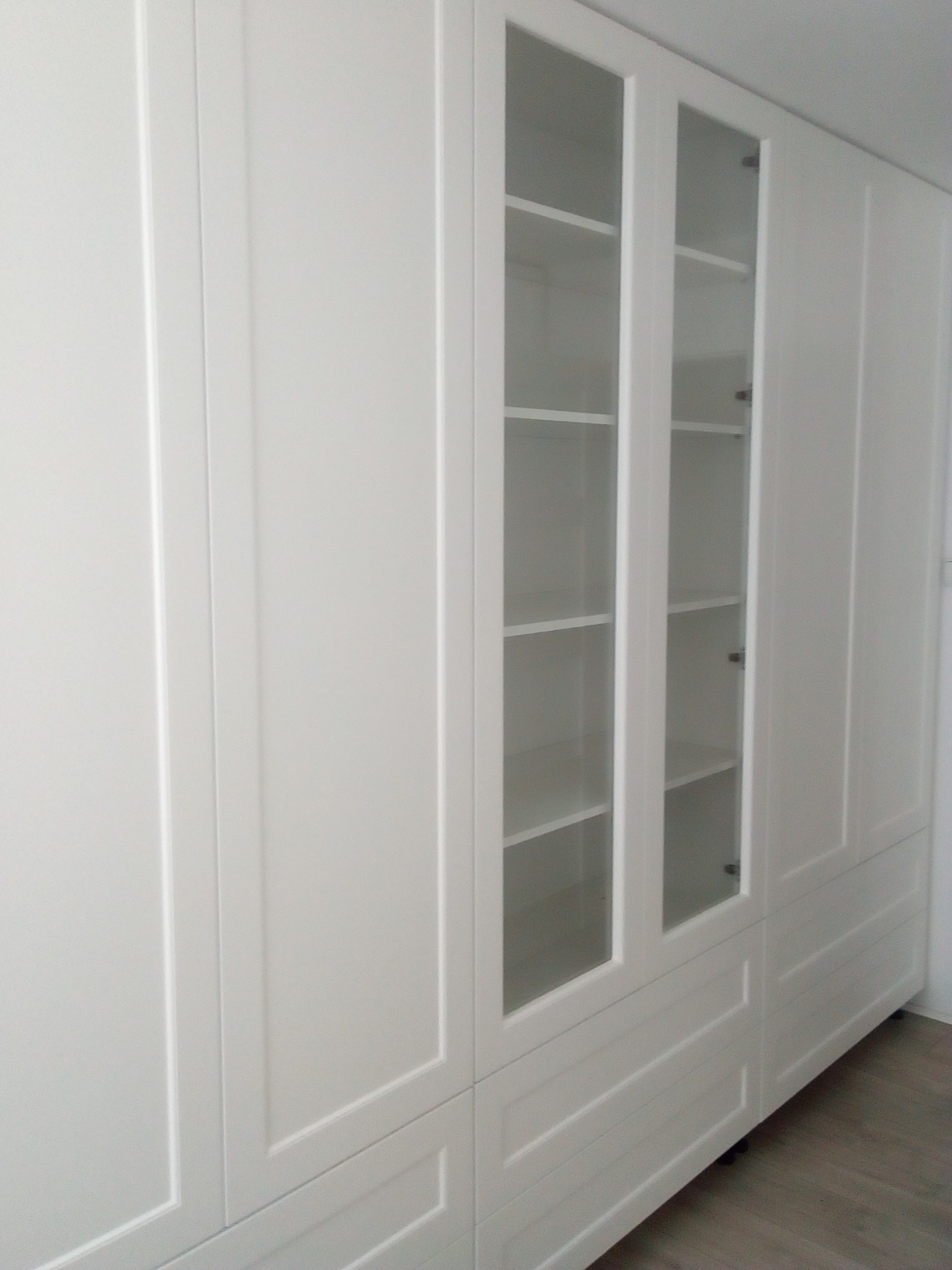 Biała szafa wnękowa z przeszklonymi drzwiami, widoczne półki i dolne szuflady, minimalistyczny design.