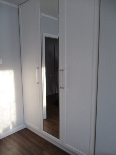 Biała szafa trzydrzwiowa z lustrem na środkowych drzwiach, widoczna podłoga z paneli i fragment ściany w neutralnym kolorze.