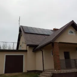 Dom jednorodzinny z brązową dachówką i panelami słonecznymi na dachu, dodatkowo antena na dachu, widok z zewnątrz w pochmurny dzień.