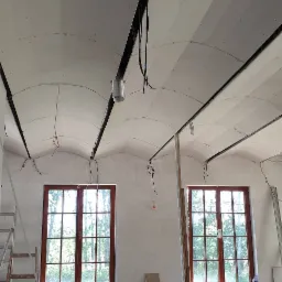Montaż płyt na łukowym suficie w pomieszczeniu z dwoma oknami, widoczne profile i instalacje elektryczne.