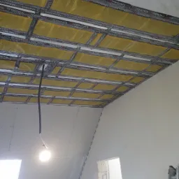 Poddasze w trakcie ocieplania, widoczne płyty kartonowo-gipsowe, folia termoizolacyjna TERMOFOL w ż&oacute;łtym kolorze, metalowy stelaż, kabel zwisający z sufitu i drabina oparta o ścianę.