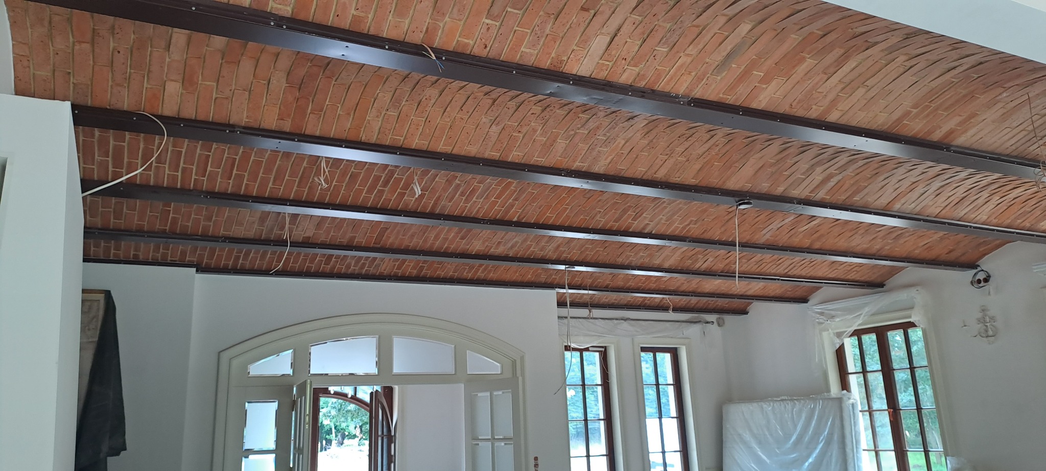 Sklepiony sufit z cegły, wzmocniony metalowymi belkami, w trakcie prac wykończeniowych w jasnym wnętrzu z oknami.