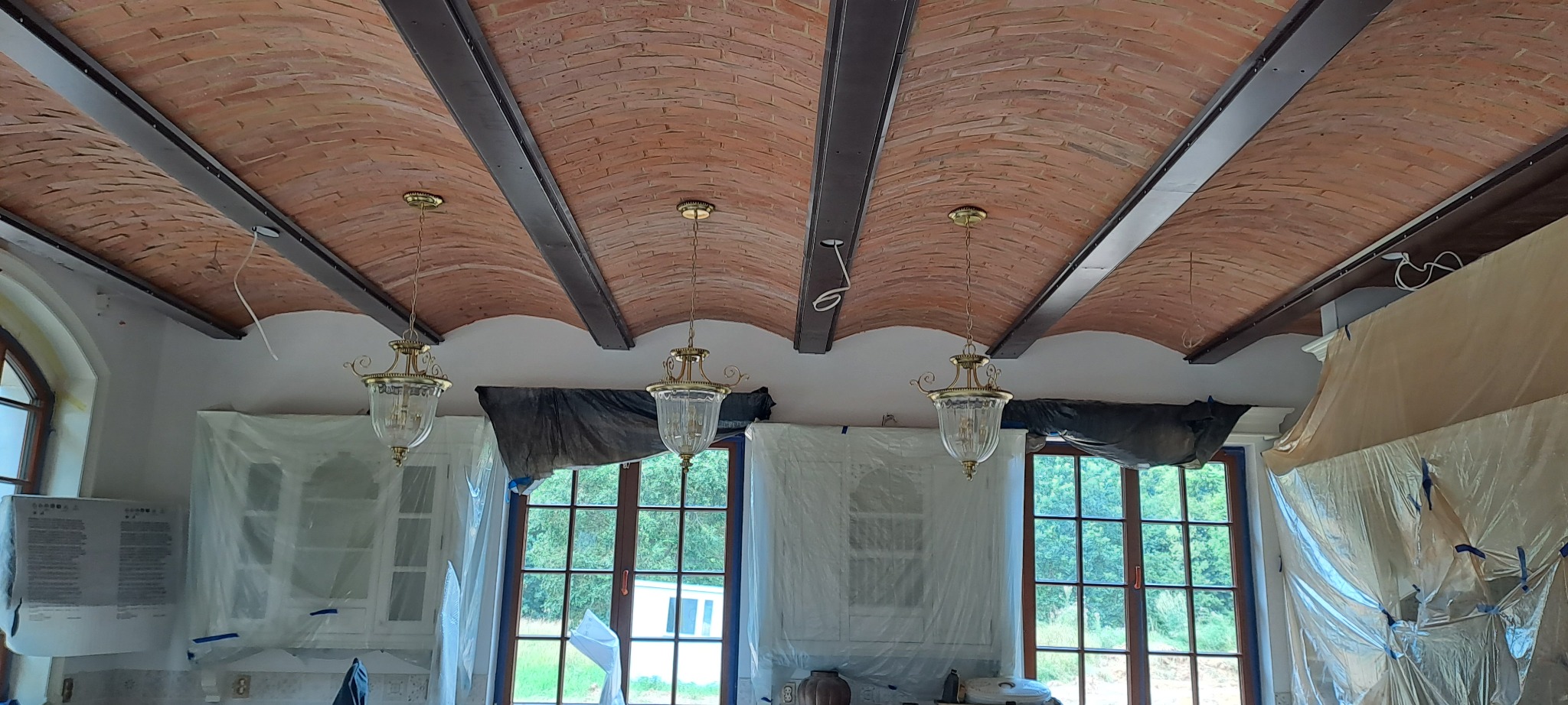Rustykalny ceglany sufit łukowy z metalowymi belkami i wiszącymi lampami w trakcie prac wykończeniowych, zabezpieczone okna i meble folią malarską.