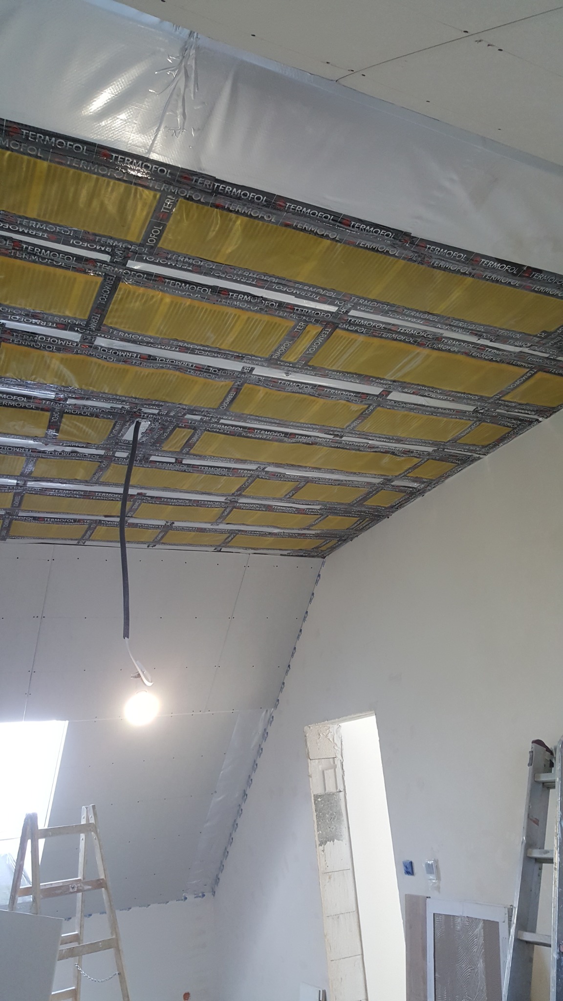 Poddasze w trakcie ocieplania, widoczne płyty kartonowo-gipsowe, folia termoizolacyjna TERMOFOL w żółtym kolorze, metalowy stelaż, kabel zwisający z sufitu i drabina oparta o ścianę.