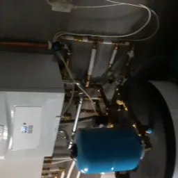 Nowoczesny piec gazowy Vaillant z podłączeniami hydraulicznymi, widoczny niebieski zbiornik wyrównawczy i miedziane rury, zamontowany na szarej ścianie.