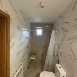 AWER-BUD - Wykończona łazienka z beżowymi płytkami imitującymi marmur, kabiną prysznicową z zasłoną, toaletą i grzejnikiem. Widoczne uchwyty dla osób z niepełnosprawnościami.