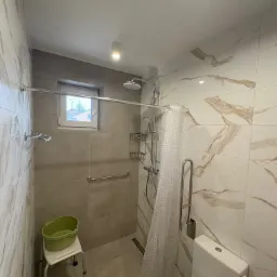 Jasna łazienka po remoncie z kabiną prysznicową, sedesem i uchwytami dla niepełnosprawnych. Marmurowe płytki na ścianach, okno w strefie prysznica.