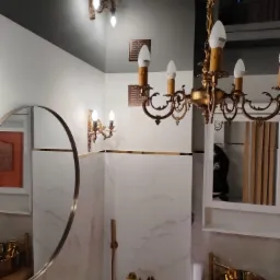 Elegancka łazienka w bieli i złocie: okrągłe lustro, marmurowe płytki, złota armatura, ozdobny żyrandol i kinkiety w stylu retro.