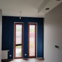 Wnętrze pokoju po remoncie: ściana pomalowana na granatowo z dekoracyjnymi panelami, drewniane ramy okienne z żaluzjami, białe ściany i sufit z oświetleniem punktowym, oraz panele podłogowe...