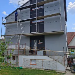 Dwukondygnacyjny dom w trakcie malowania elewacji na szaro, z fragmentem pomalowanej czarnej ściany, rusztowania wokół budynku, pracownik na rusztowaniu z puszką farby, zabezpieczone okna folią.