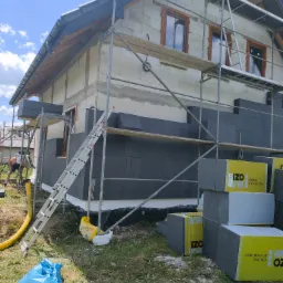 Ocieplanie elewacji domu jednorodzinnego z użyciem styropianu grafitowego, widoczne rusztowanie, niebieski worek na śmieci i opakowania styropianu IZO Line