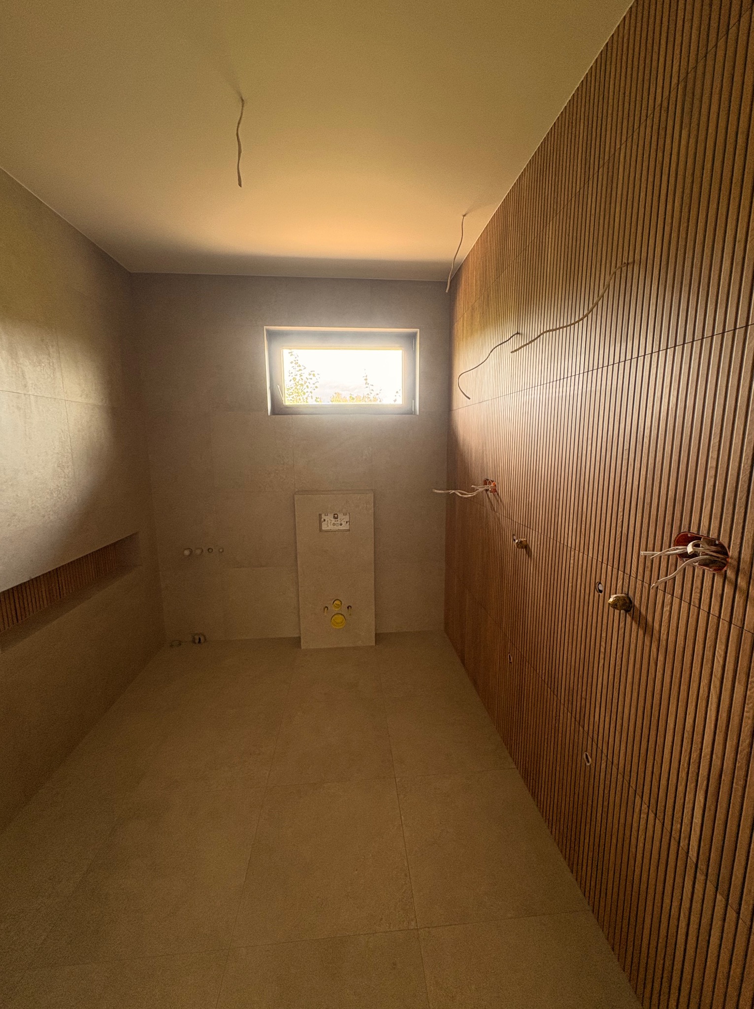 Wnętrze łazienki w trakcie wykończenia: płytki na podłodze i ścianach, drewniana boazeria, okno i instalacja sanitarna. Widoczne przewody elektryczne.