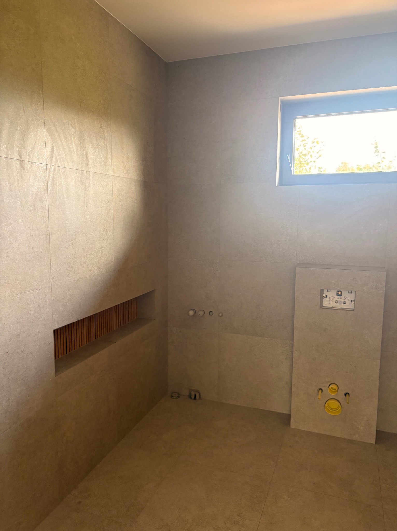 Wykończona łazienka z beżowymi płytkami, wnęką z drewnianymi listwami i oknem. Widoczne instalacje podtynkowe i stelaż WC. Minimalistyczny design wnętrza.