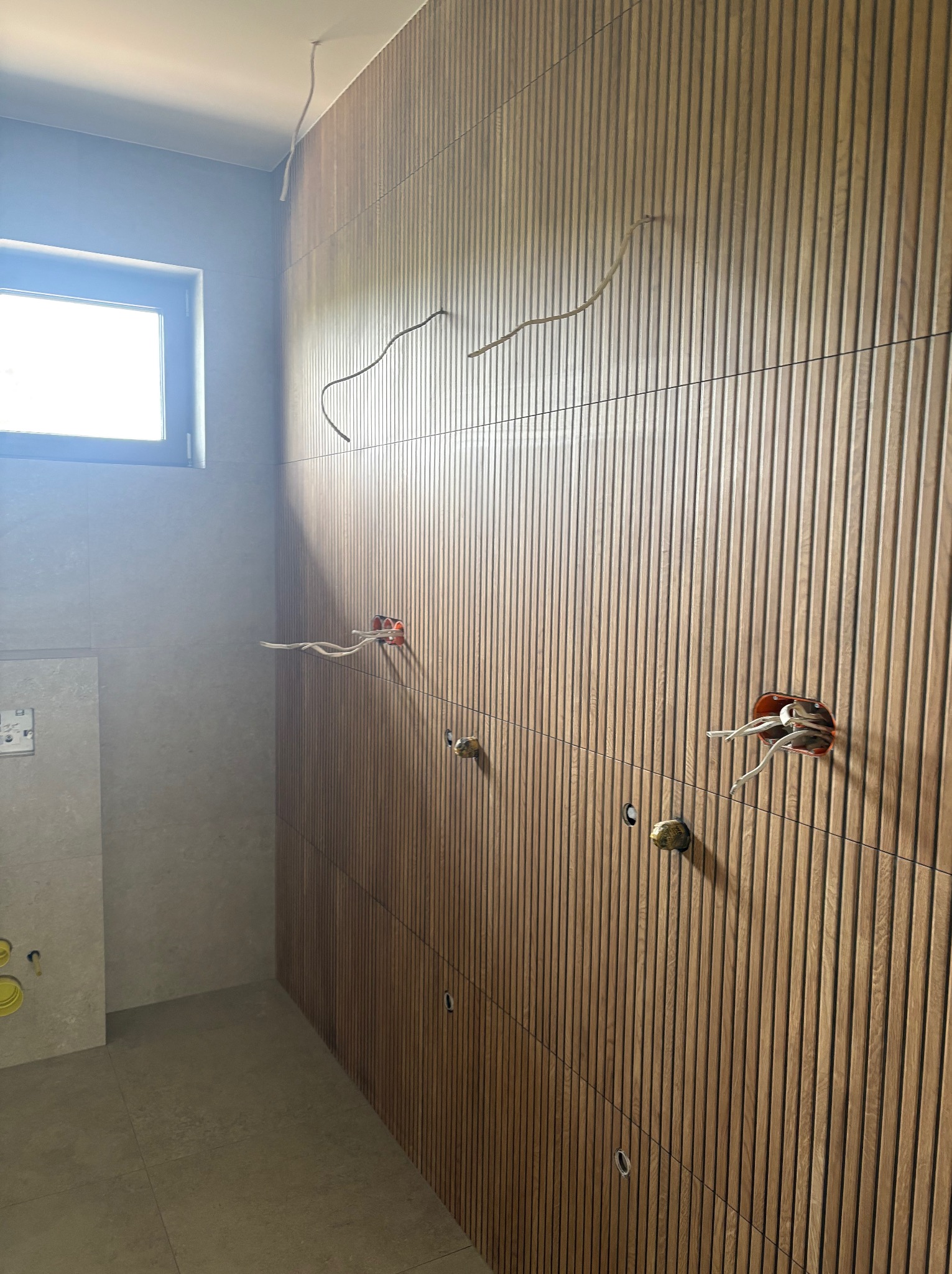 Łazienka w trakcie remontu: drewniana ściana z pionowymi panelami, widoczne puszki elektryczne i wystające przewody. Na ścianie obok szare płytki i okno.
