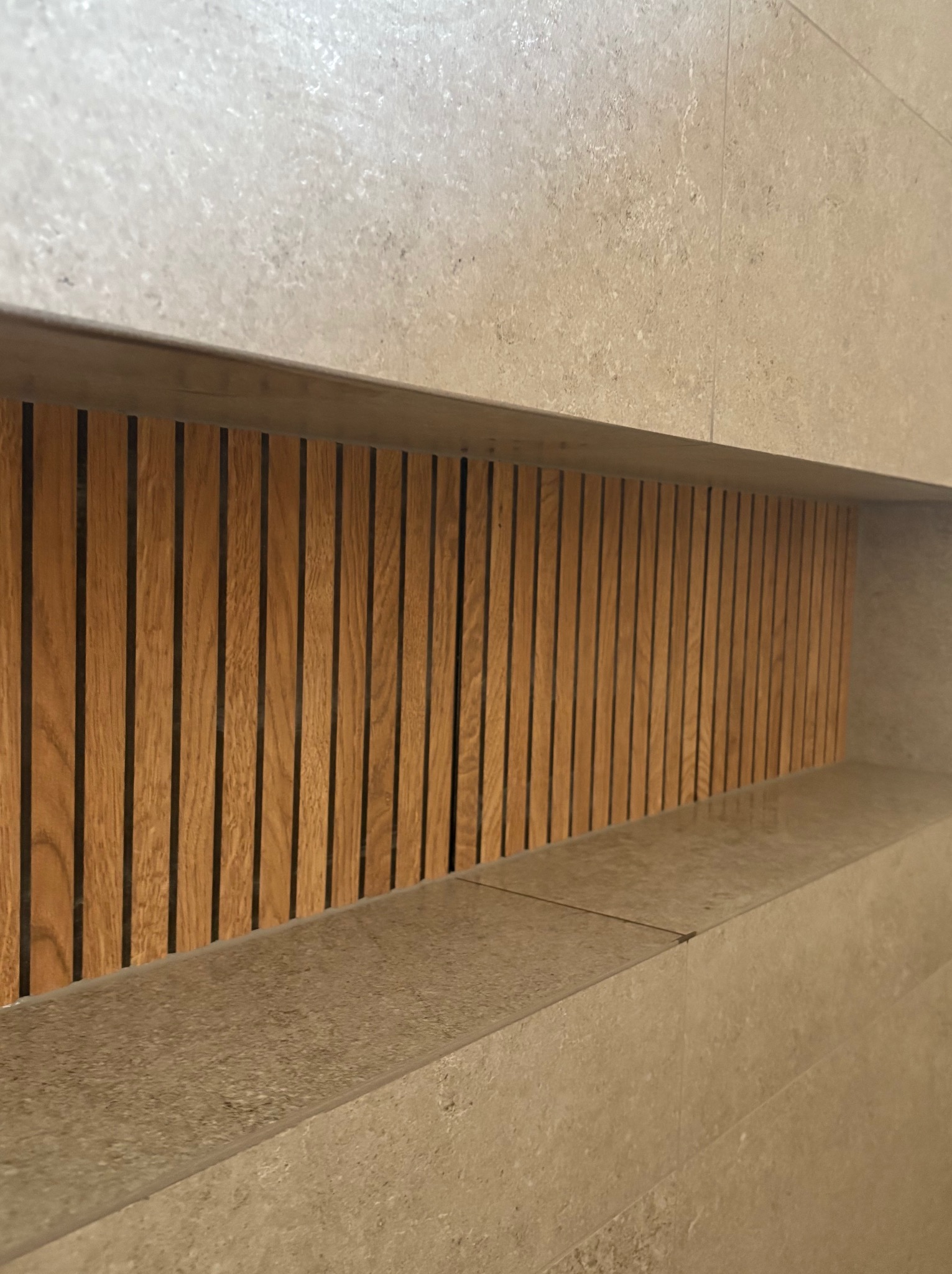 Narożne ujęcie wnęki ściennej wyłożonej beżowymi płytkami, z dekoracyjnymi, pionowymi listwami z jasnego drewna wewnątrz. Minimalistyczny design.