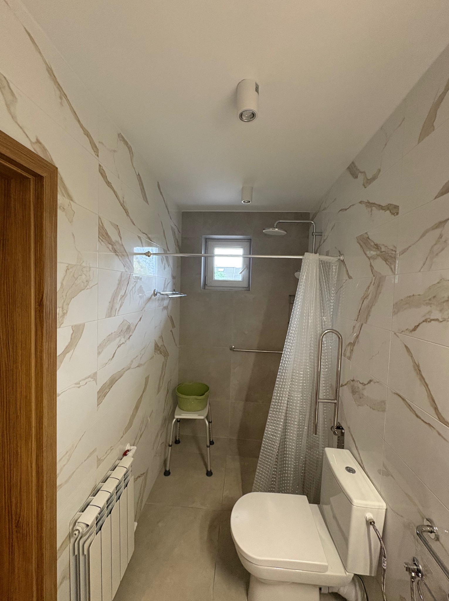 Wykończona łazienka z beżowymi płytkami imitującymi marmur, kabiną prysznicową z zasłoną, toaletą i grzejnikiem. Widoczne uchwyty dla osób z niepełnosprawnościami.