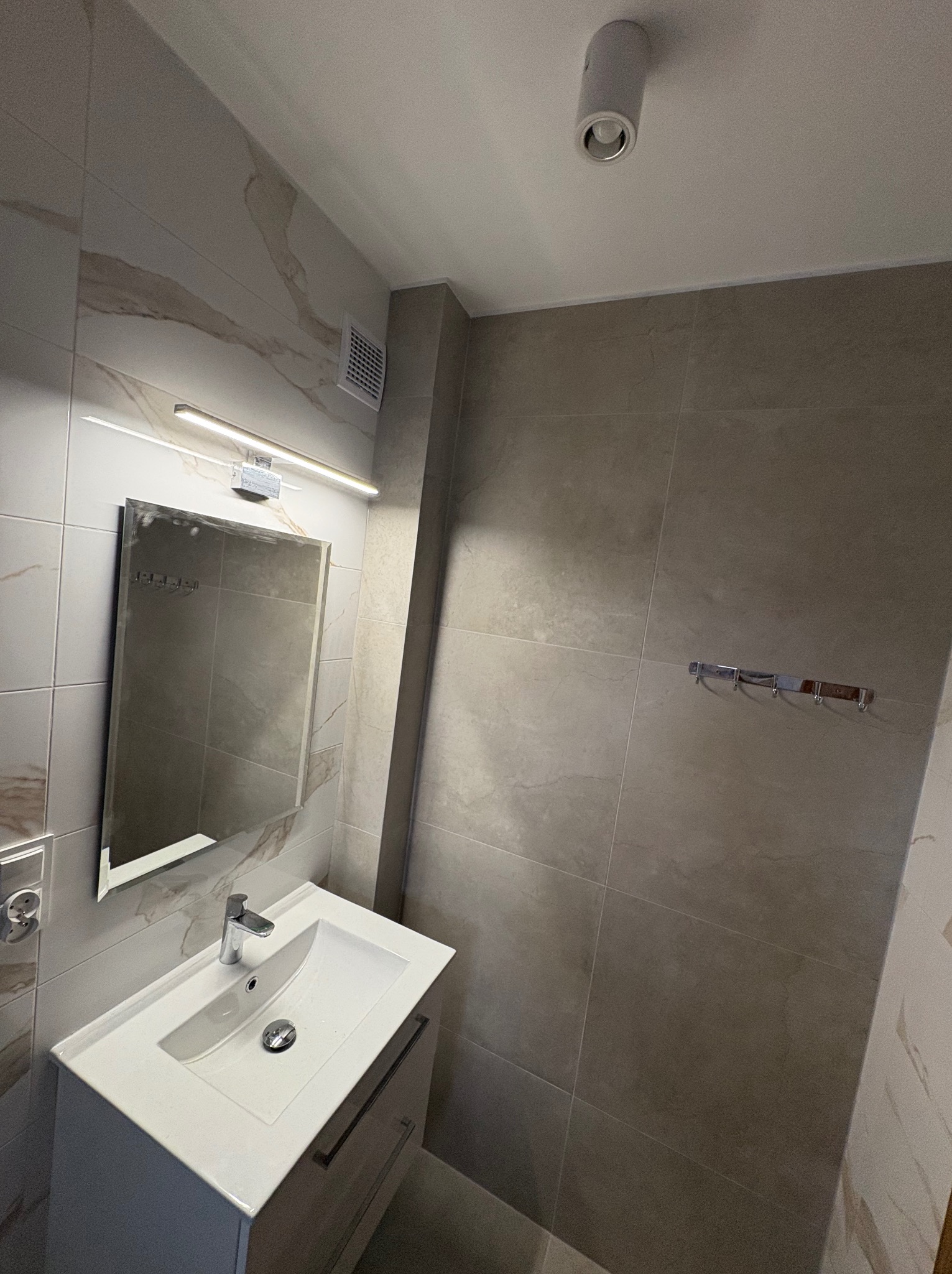 Nowoczesna łazienka z beżowymi płytkami imitującymi marmur, białą umywalką z szafką i lustrem podświetlonym listwą LED. Widoczny wieszak na ręczniki i punktowe oświetlenie.