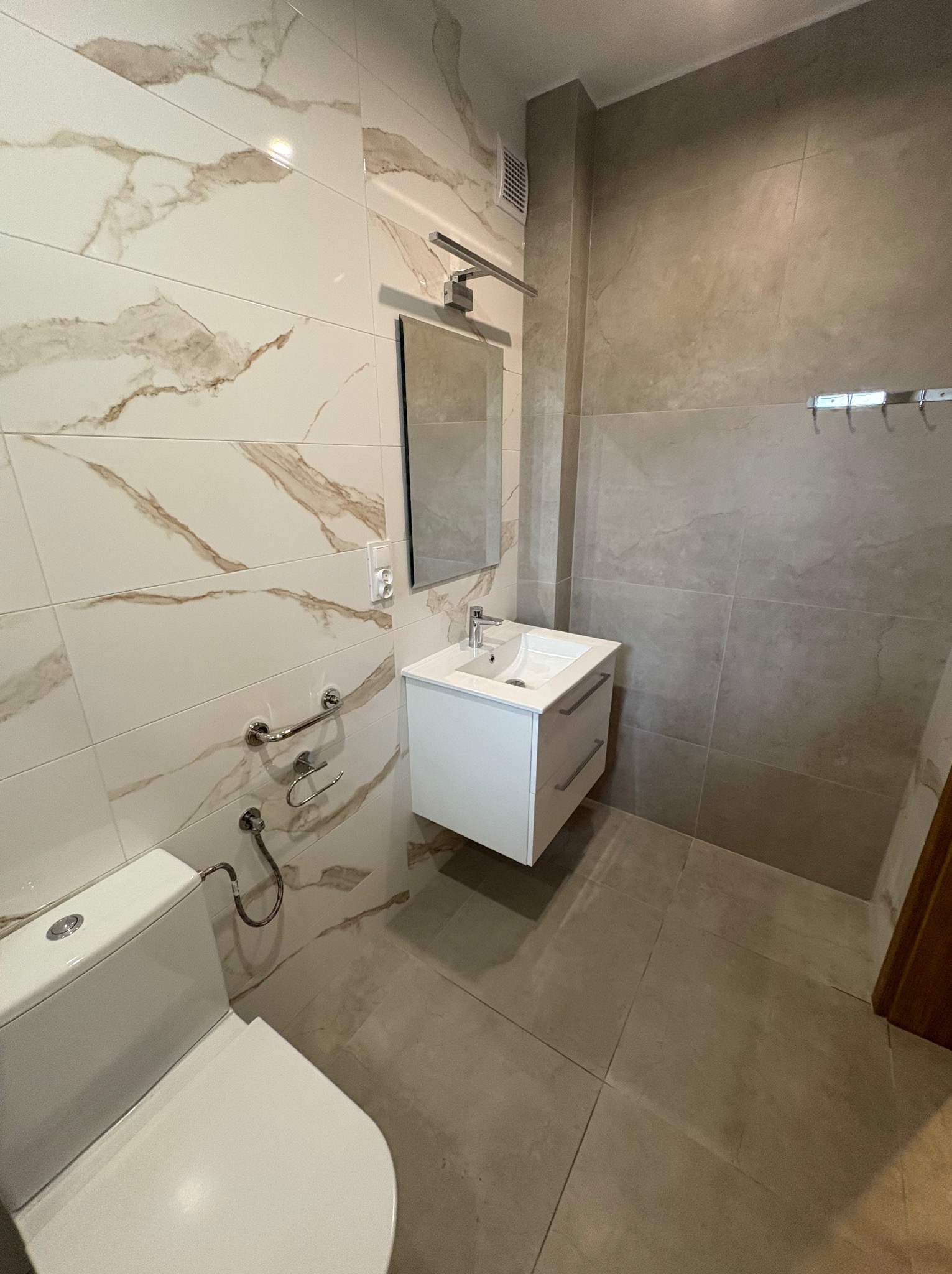 Wykończona łazienka z płytkami imitującymi marmur na ścianach, białą szafką umywalkową i wiszącą toaletą. Widoczne detale armatury i oświetlenia.