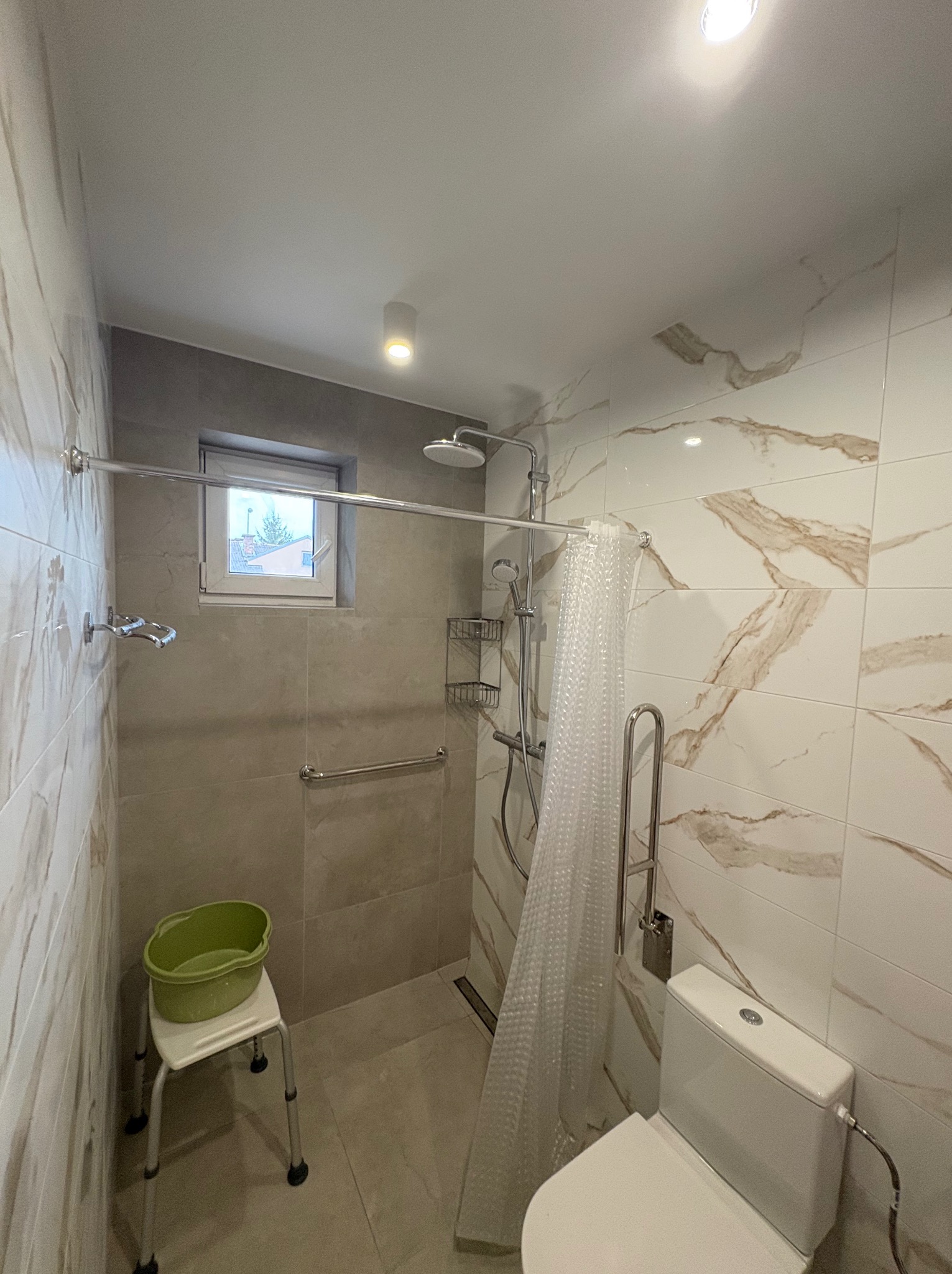 Jasna łazienka po remoncie z kabiną prysznicową, sedesem i uchwytami dla niepełnosprawnych. Marmurowe płytki na ścianach, okno w strefie prysznica.