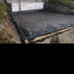 Przygotowanie zbrojenia na deskowaniu pod płytę fundamentową, widoczne zabezpieczenie folią, wykop ziemny i fragment budynku w tle.