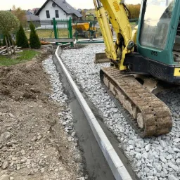 Żółta mini koparka obok ułożonego krawężnika w trakcie prac ziemnych, widoczne przygotowane podłoże z kamieni i ziemi, w tle dom jednorodzinny i zielone tuje.