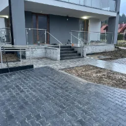 Nowoczesny dom z szarą elewacją, szklanymi balustradami na balkonie i tarasie, schody z ciemnego kamienia i świeżo ułożona, wilgotna kostka brukowa w odcieniach szarości.