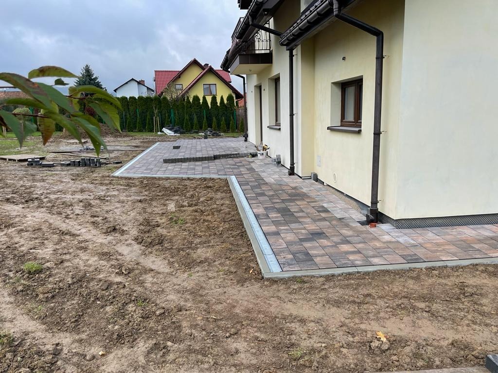 Częściowo ułożony chodnik z kostki brukowej w odcieniach brązu i szarości, z widocznym systemem odprowadzania wody, przylegający do jasnej elewacji domu jednorodzinnego z brązową stolarką okienną...