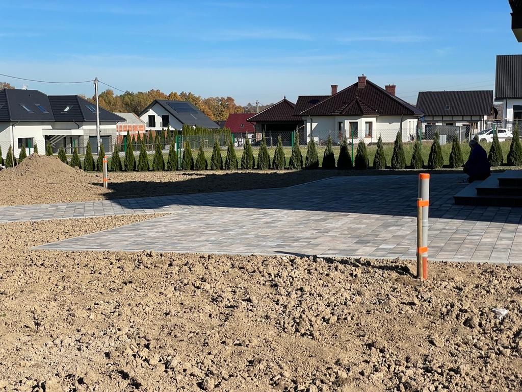 Fragment posesji z ułożoną szarą kostką brukową w geometryczny wzór, widoczny stos ziemi, rząd małych drzewek iglastych i domy jednorodzinne w tle, słoneczny dzień.