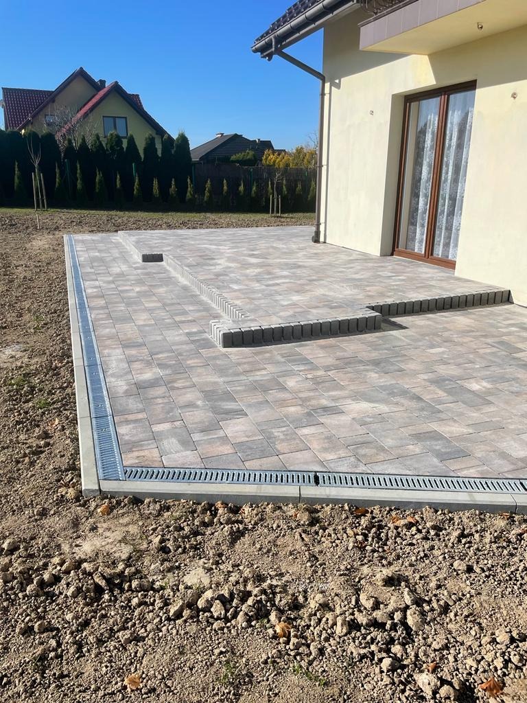 Nowoczesny taras z kostki brukowej w odcieniach szarości, z liniowym odwodnieniem, prowadzący do budynku z brązowymi oknami, na tle zieleni i błękitnego nieba.