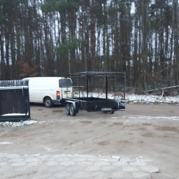 Filtum - Czarna, metalowa przyczepka samochodowa z klatką, zaparkowana na błotnistym placu w zimowy dzień, widoczny biały samochód dostawczy i las w tle.