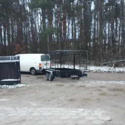 Czarna, metalowa przyczepka samochodowa z klatką, zaparkowana na błotnistym placu w zimowy dzień, widoczny biały samochód dostawczy i las w tle.