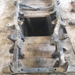 Filtum - Metalowa rama pojazdu po demontażu, widoczne ślady korozji i zabrudzenia, ułożona na drewnianej podłodze warsztatu.