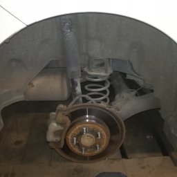 Filtum - Widok na tylne zawieszenie samochodu z widoczną tarczą hamulcową, sprężyną i amortyzatorem, zardzewiałe śruby mocujące koło.